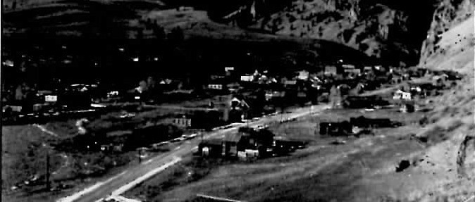 Black-and-white aerial view of the mining town of Creede, Colorado, its buildings and dirt streets strung along a narrow valley floor between steep, sparsely wooded mountainsides.