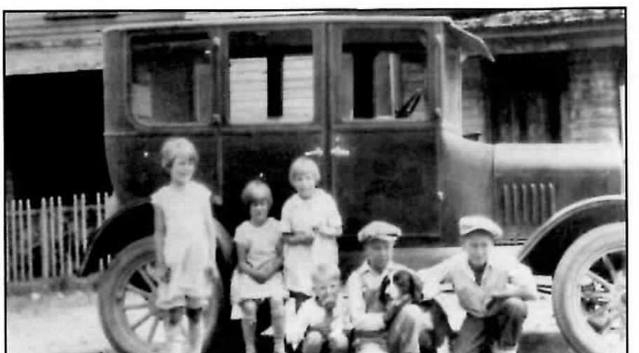 Black-and-white photograph of six children posed in front of a parked automobile outside a wood-frame building, three girls standing at left in light dresses and three boys sitting or crouching at right in caps and work clothes, one boy holding a dog.