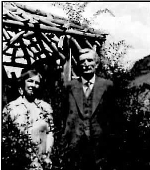 Black-and-white photograph of an older man in a suit and a woman in a patterned dress standing together outdoors in front of a rustic wood arbor, with mountains in the background.