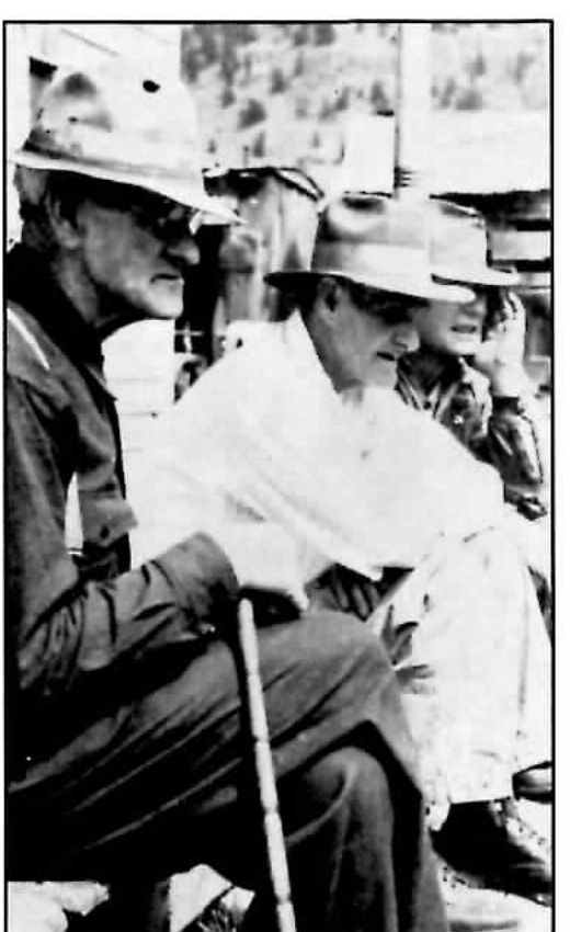 Black-and-white photograph of three older men seated closely together outdoors, the nearest wearing a broad-brimmed hat and holding a cane, the middle man in a light-colored jacket and hat, the third partly turned away.