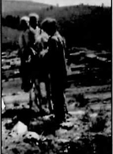 Black-and-white photograph of three boys standing beside a burro on an open, sloping hillside, with a scattering of small buildings visible in the valley behind them.