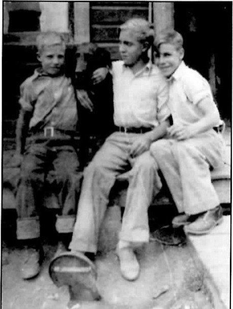 Black-and-white photograph of three boys standing and sitting together outdoors in front of a wood building, the tallest with an arm around the shoulders of the boy beside him, all three in everyday work clothes and boots.
