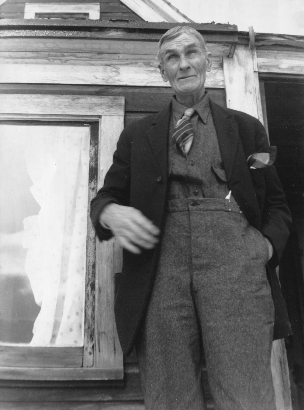 Black-and-white photograph, taken from a low angle, of an elderly man in a dark suit coat, wool vest, patterned tie, and pocket handkerchief, one hand in his pocket, standing beside a curtained window and an open doorway of a weathered wood-frame house with a gable above.