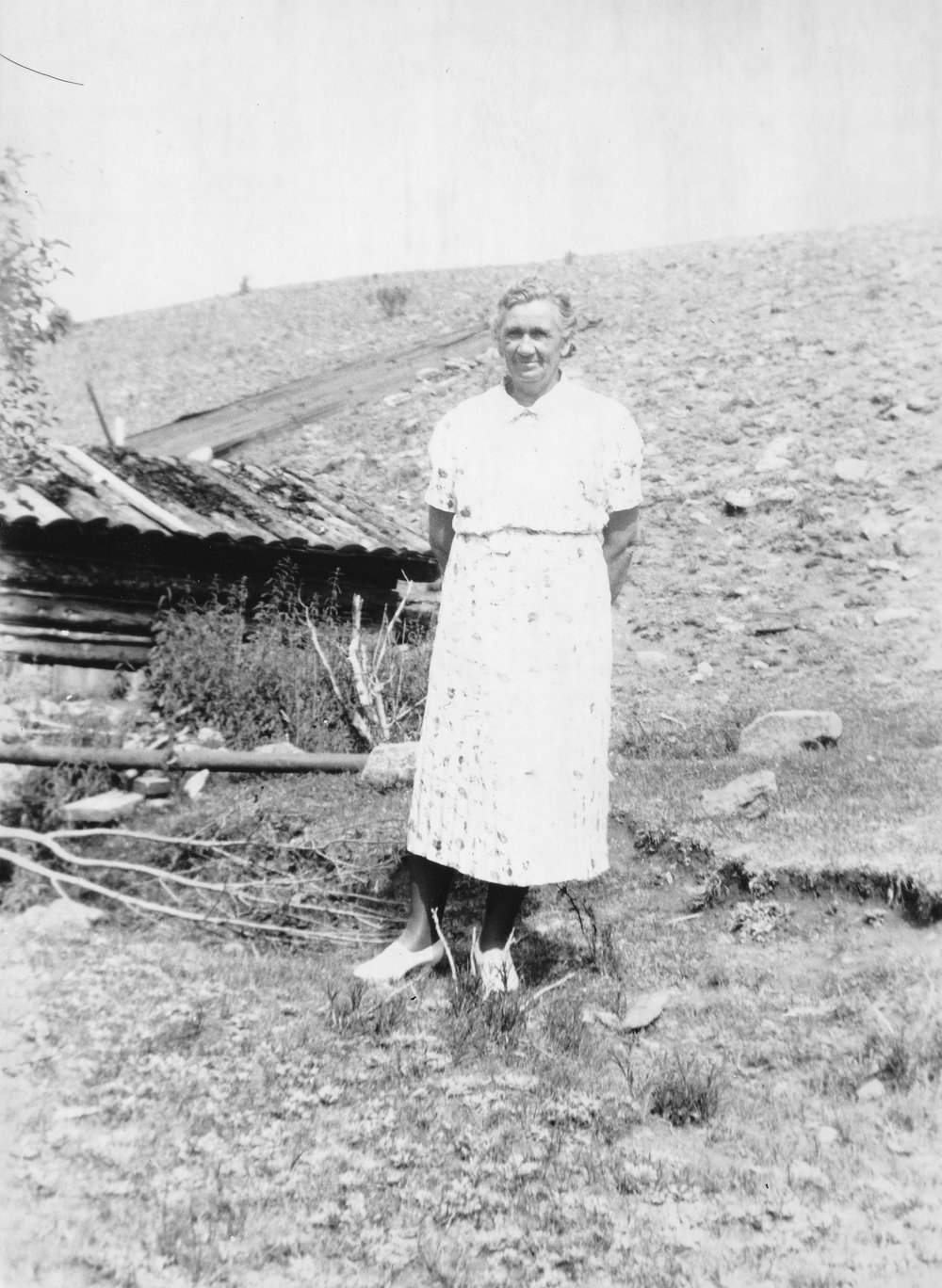 Black-and-white photograph of an older woman with gray hair, in a light print dress and white shoes, standing with her hands behind her back in front of a low log structure with a sagging, partly broken roof, set against a steep, rocky hillside.