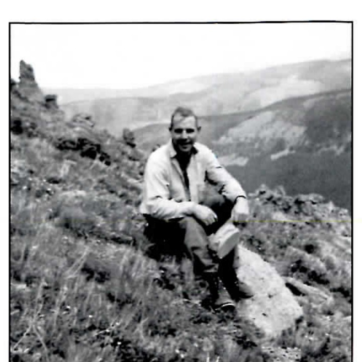 Black and white photograph of a man sitting on a rocky hillside overlooking a mountain valley.