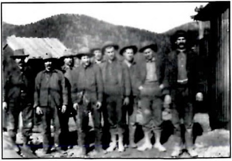 Black-and-white photograph of a row of about a dozen men in work clothes and hats standing in front of a timber building with a forested hill behind them.