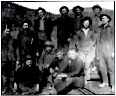 Black-and-white group photograph of about a dozen men in work clothes and hats, some standing and some crouching, in front of a forested hillside.