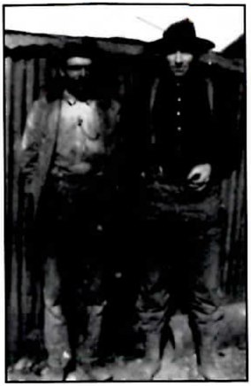 Black-and-white photograph of two men standing side by side against a wall of corrugated metal: on the left a bearded man in a light, open coat, and on the right a taller man in a dark hat and buttoned dark jacket.
