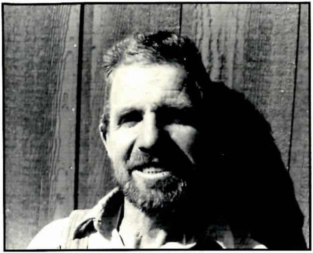 Black-and-white portrait of a smiling bearded man with short hair, in a collared shirt and vest, photographed against a wall of vertical wooden boards with deep shadow falling across the right side of the frame.