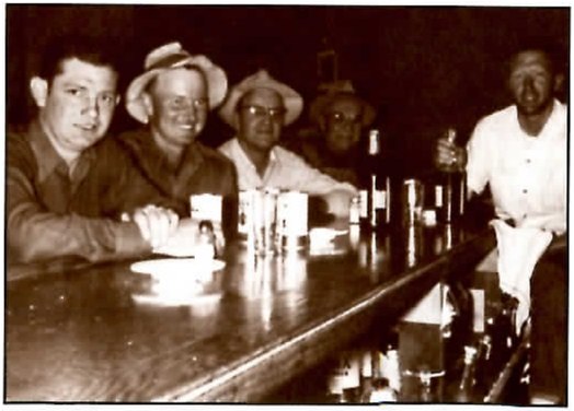 Sepia photograph of five men along a bar, taken from the end of the counter: three seated men in hats or work shirts face the camera, a fourth sits in shadow behind them, and a bartender in a white shirt stands at right holding a bottle. Glasses, a napkin holder and bottles crowd the bar top.