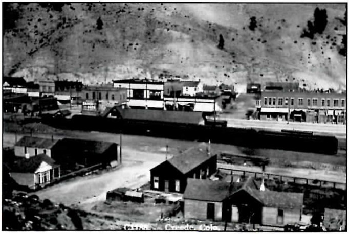 Black-and-white view down onto the main street and railroad depot of Creede, Colorado, with a long dark passenger train standing along the tracks below a bare, scattered-brush hillside. Part of a printed line along the bottom edge reads "Creede, Colo."