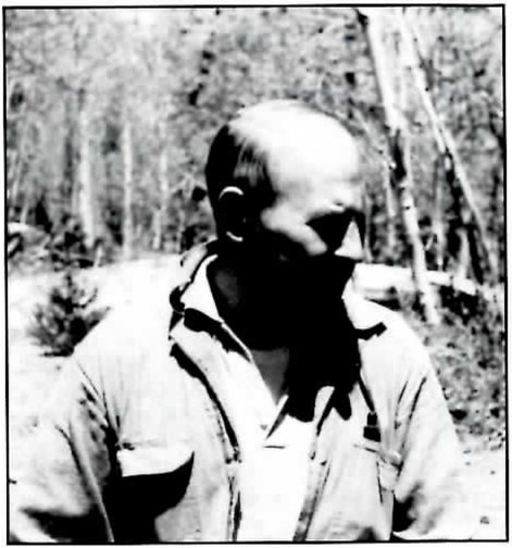 Black-and-white photograph of a balding man in a short-sleeved collared shirt, seen from the chest up and looking off to one side, with trees behind him.