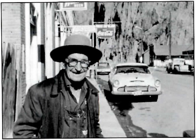 Black-and-white photograph of a smiling older man in a dark brimmed hat, glasses, a jacket and bib overalls, standing on a sidewalk in a small-town street, with a parked 1950s car, a Coca-Cola sign and other buildings behind him.