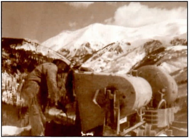 Sepia photograph of a man bent over a large cylindrical ore car, with a snow-covered mountain and clouds behind it.