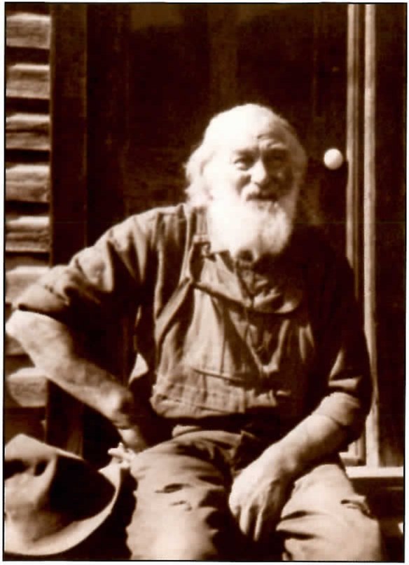 Sepia photograph of an elderly man with white hair and a full white beard, in a work shirt and overalls, sitting in a log-cabin doorway with a hat beside him and smiling toward the camera.