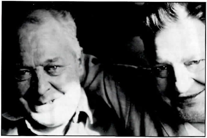 Black-and-white close-up of two men's faces: at left an older man with white hair and a broad smile, and at right a younger man with dark hair leaning in beside him.