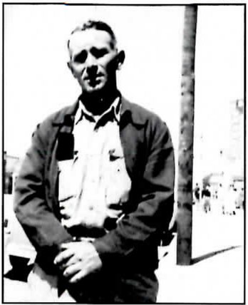 Black-and-white photograph of a man in a dark work jacket over an open-collared shirt, standing with his hands clasped in front of him beside a utility pole on a town street, with buildings and parked cars blurred behind him.