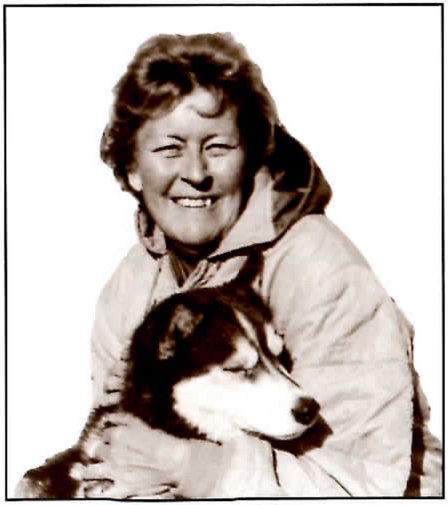 Sepia photograph of a smiling woman with short curly hair, wearing a quilted hooded jacket, hugging a husky-type dog with a dark-and-white face against her chest.