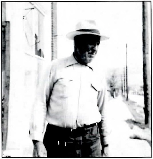 Black-and-white photograph of a heavyset man in a light hat and shirt standing on a sidewalk beside a building.