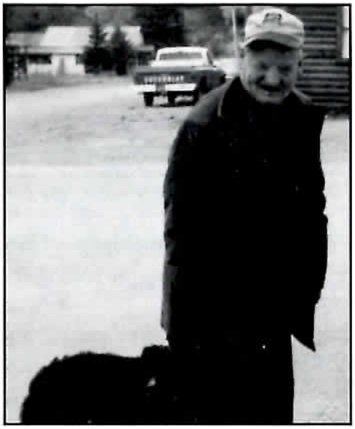 Black-and-white photograph of an older man in a ball cap and dark jacket, smiling as he looks down, standing on a gravel street with a pickup truck and buildings behind him and a dark dog in the lower left foreground.