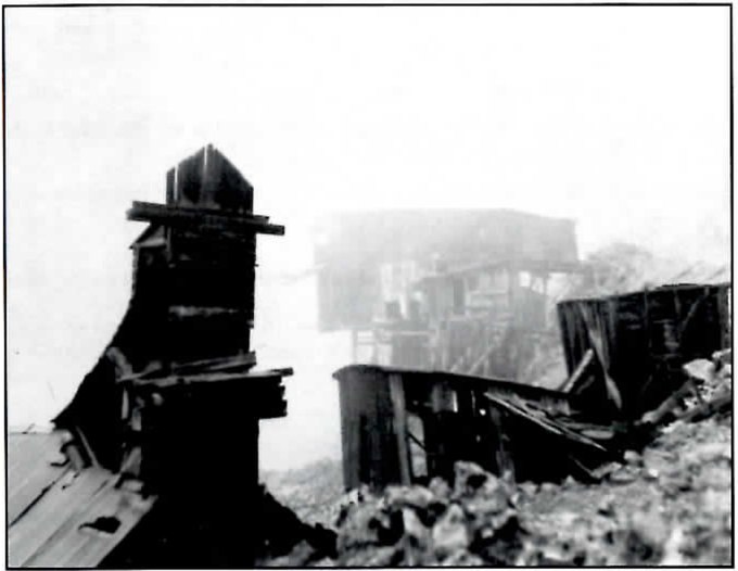 Black-and-white photograph of collapsed timber mine buildings and a tall, chimney-shaped wooden structure in the foreground, with more weathered buildings fading into fog behind them.