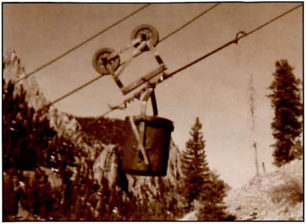 Sepia photograph of an ore bucket hanging from a pulley carriage on aerial tram cables above a rocky, timbered canyon.