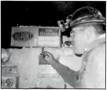 Black-and-white photograph taken underground of a man in profile, in glasses and a hard hat with a headlamp, pointing with one raised hand at a box marked DuPont High Explosives.