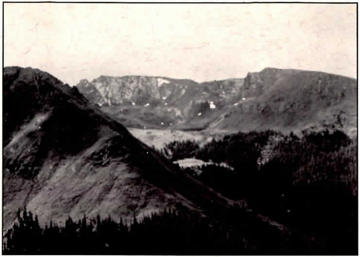 Black-and-white photograph of a high mountain basin ringed by rocky, snow-streaked peaks, with dark timber on the near slope and a pale meadow in the valley beyond.