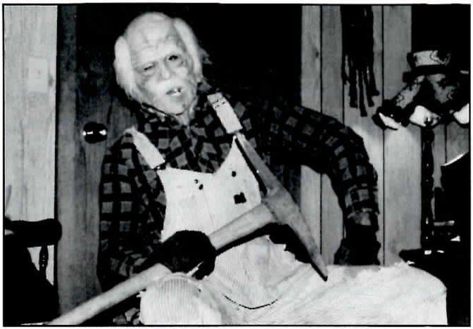 Black-and-white photograph of a white-haired figure with a lined face and few teeth, seated in a checked flannel shirt, white bib overalls and dark gloves, holding a pick across the lap, in a paneled room with a hat and other objects hanging on the wall behind.