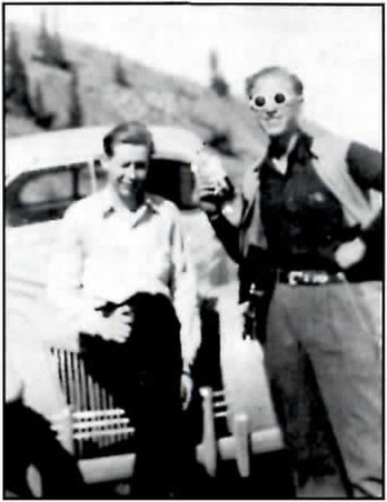 Black-and-white photograph of two men beside a 1930s car: a young man in a light shirt sits on the front fender at left, and a taller man in a vest and round, white-rimmed novelty glasses stands beside him with one hand on his hip.