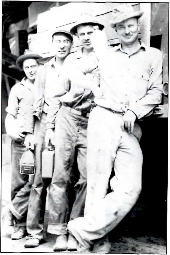Black-and-white photograph of four men in work clothes and hats standing close together in front of a wooden wall; the two at left hold lunch pails, and the mustached man at right leans back with one hand on his hip.