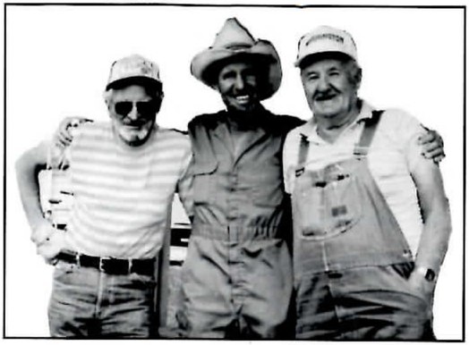 Black-and-white photograph of three smiling men with their arms around one another's shoulders: at left a bearded man in sunglasses, a cap and a striped shirt; in the middle a man in a cowboy hat and coveralls; at right a mustached man in a ball cap and bib overalls.