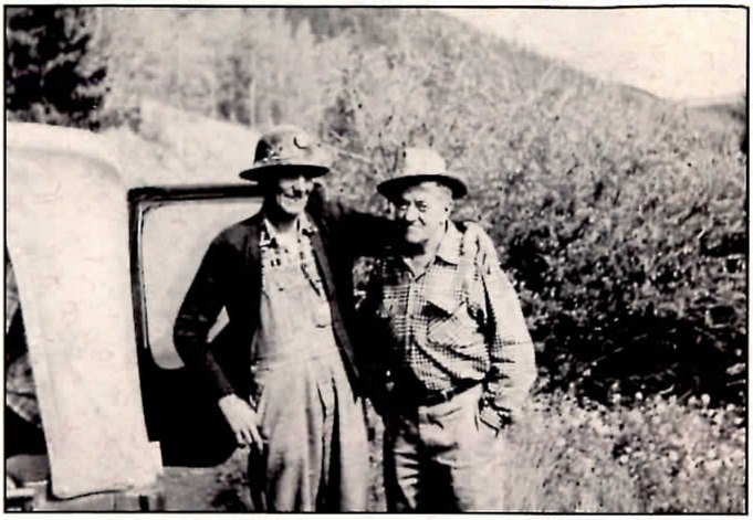 Black-and-white photograph of two men in hats standing side by side with their arms around each other, beside the open door of an old vehicle, in front of a brushy hillside. The man at left wears a miner's hat with a lamp, a dark cardigan and overalls; the man at right wears a plaid work shirt.