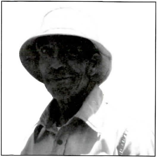 Black-and-white close photograph, heavily contrasted, of a man in a light bucket hat and a collared work shirt, smiling slightly, his face in shadow under the brim.