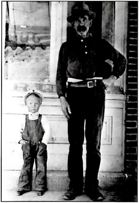 Black-and-white photograph of a tall man with a mustache, in a dark shirt, belt and brimmed hat, standing with one hand on his hip on a storefront doorstep beside a small boy in overalls and a cap.