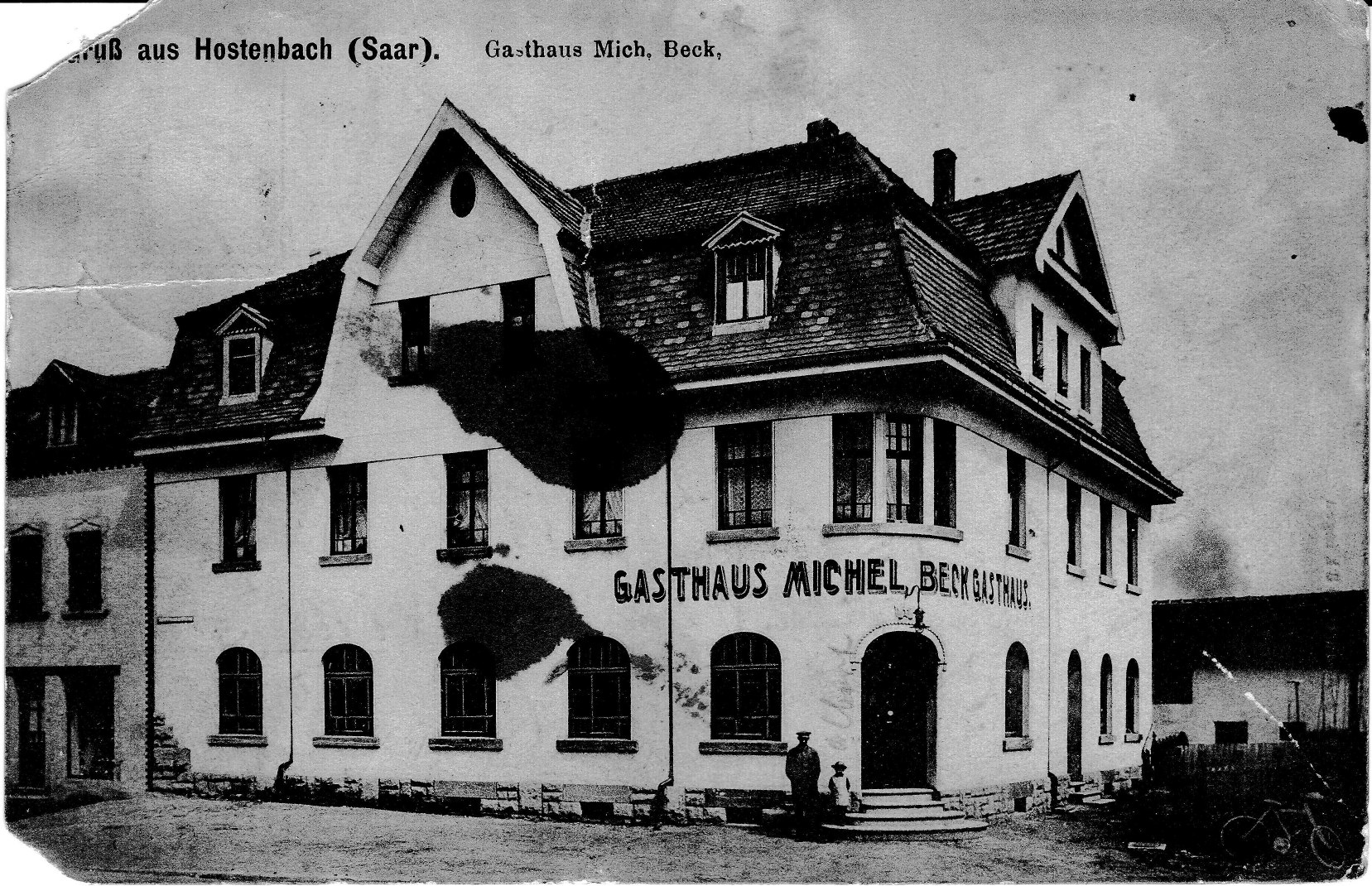 Black-and-white photograph postcard of a large, newly built two-and-a-half-story inn with a steep tiled roof, dormers, and arched ground-floor windows. Lettering across the facade reads Gasthaus Michel Beck Gasthaus. A man and a small child stand by the arched front door. The printed heading reads Gruß aus Hostenbach (Saar), Gasthaus Mich. Beck. Dark water stains mark the upper left of the image.